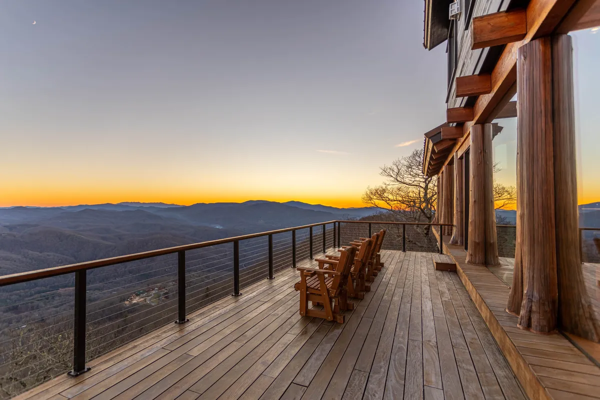 A pair of wooden chairs are positioned on a deck facing out towards a mountain vista, with a sunset sky overhead. The cozy arrangement suggests a setting for deep conversation, stargazing, or simply enjoying the serenity of nature.