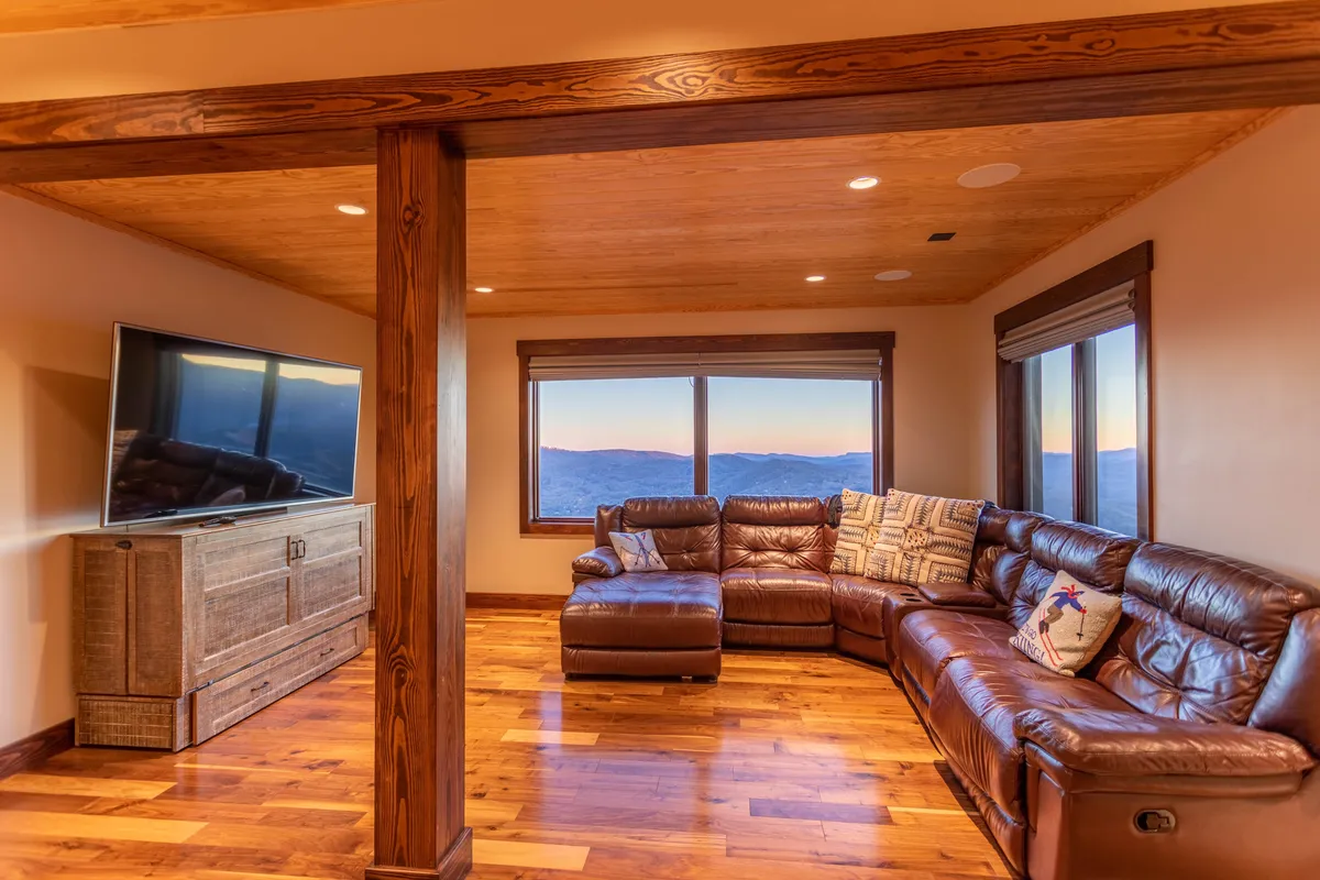 The scene captures a leather sectional couch positioned in front of a large television, with windows presenting expansive mountain views. The room is set up for movie watching and gatherings, evoking warmth and togetherness.