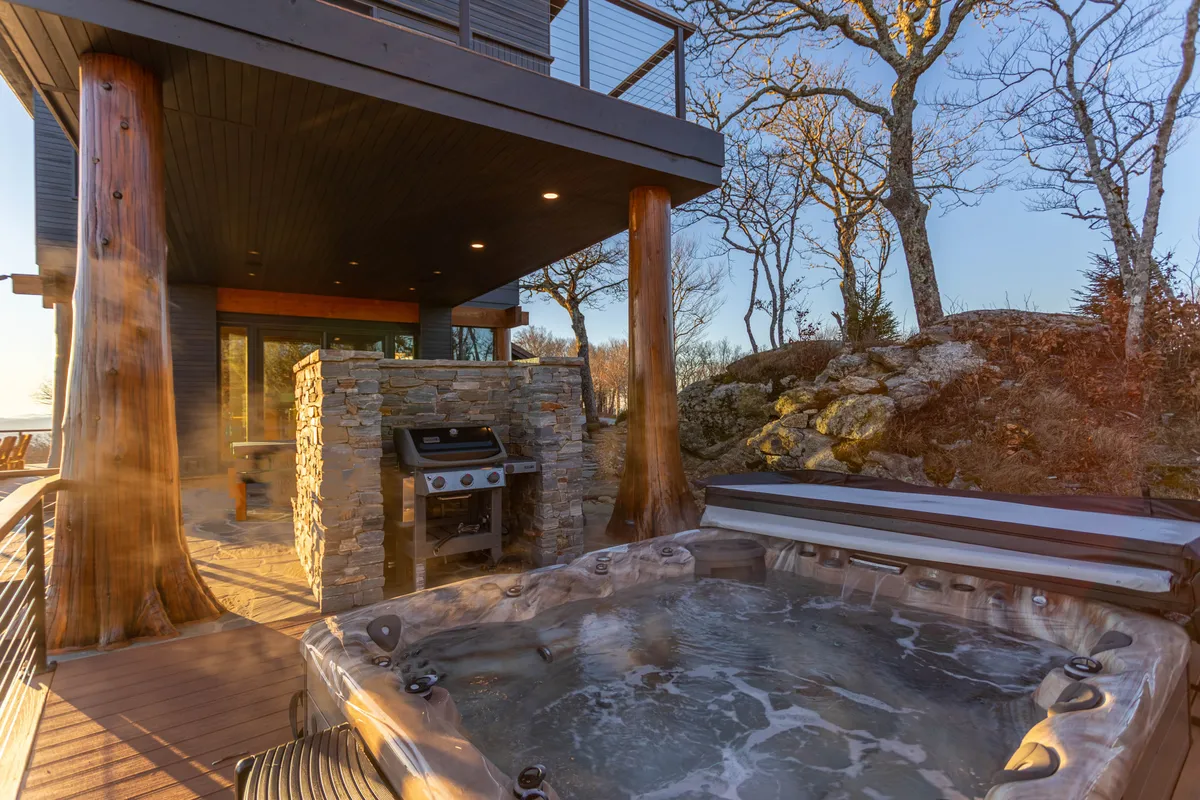 An inviting exterior shot featuring a bubbling hot tub, outdoor grill, wooden pillars, and a stone wall. The area is set against a backdrop of lush natural surroundings, perfect for relaxing after a day of adventure, with the suggestion of sunset lighting for added warmth.