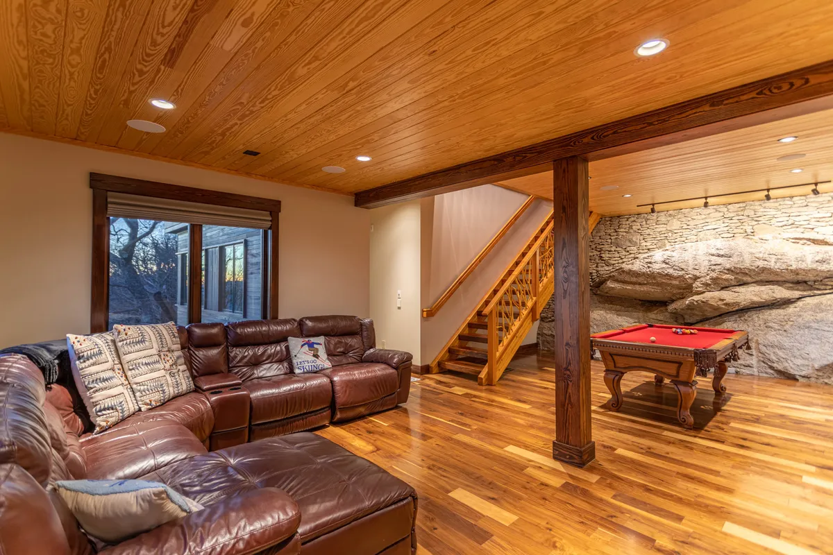 This interior image features a leather sectional sofa and a pool table set against a natural stone wall, with a staircase visible to one side. The space feels open, inviting, and designed for social activity, with furniture arranged for both conversation and gameplay.