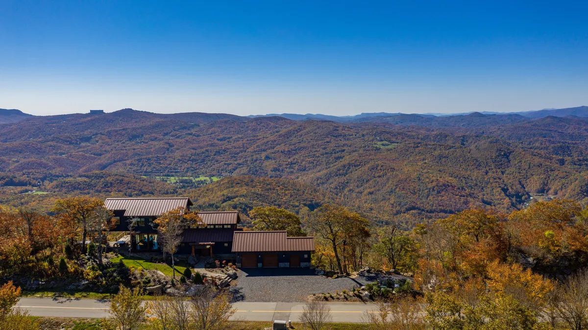 A modern home nestled among mountains with golden fall foliage, a wide driveway, and expansive outdoor areas. The environment suggests ample space for hiking, gathering, and enjoying the crisp mountain air, reinforcing the sense of an upscale basecamp for adventure.