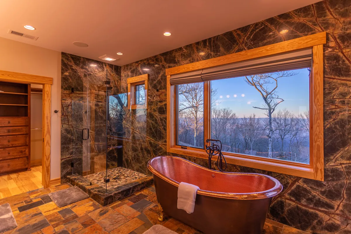 A luxurious bathroom with a striking freestanding copper soaking tub positioned near a large window that frames scenic views. The space also features a glass-enclosed rain-style shower, creating a spa-like experience bathed in natural light.