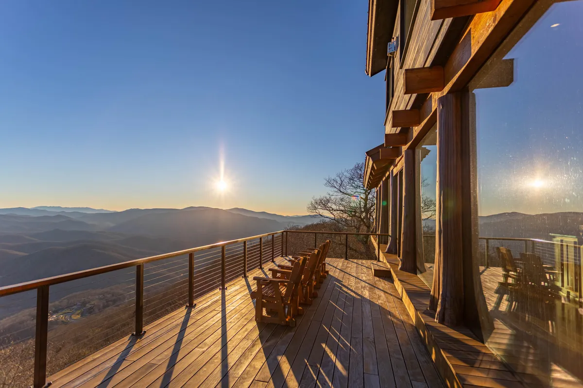 A wide wooden deck with comfortable seating overlooks a majestic mountain range at sunset. Several wooden chairs are arranged for relaxing, with panoramic views stretching into the distance. The tranquil mood suggests an ideal spot for quiet mornings, social gatherings, or stargazing.