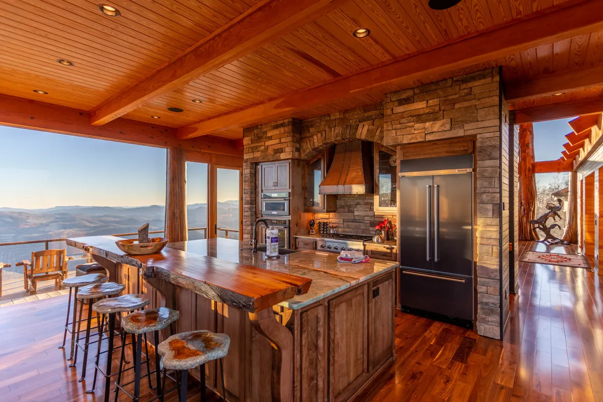 The gourmet kitchen is front and center in this image. A strikingly large, wooden kitchen island provides ample preparation and gathering space, while a stone feature wall and open sightlines lead directly to expansive views of the landscape. The design seamlessly blends social cooking, dining, and breathtaking scenery.