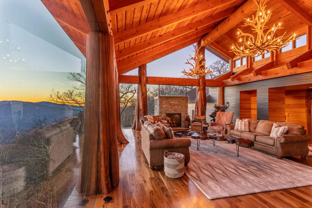 This image depicts a sunlit living room with strikingly large windows that frame breathtaking mountain views. The space features plush seating arranged around an elegant fireplace, rustic wooden beams overhead, and an antler chandelier, exuding a sense of comfort and luxury. The open layout signals a room designed for both intimate gatherings and lively family moments.