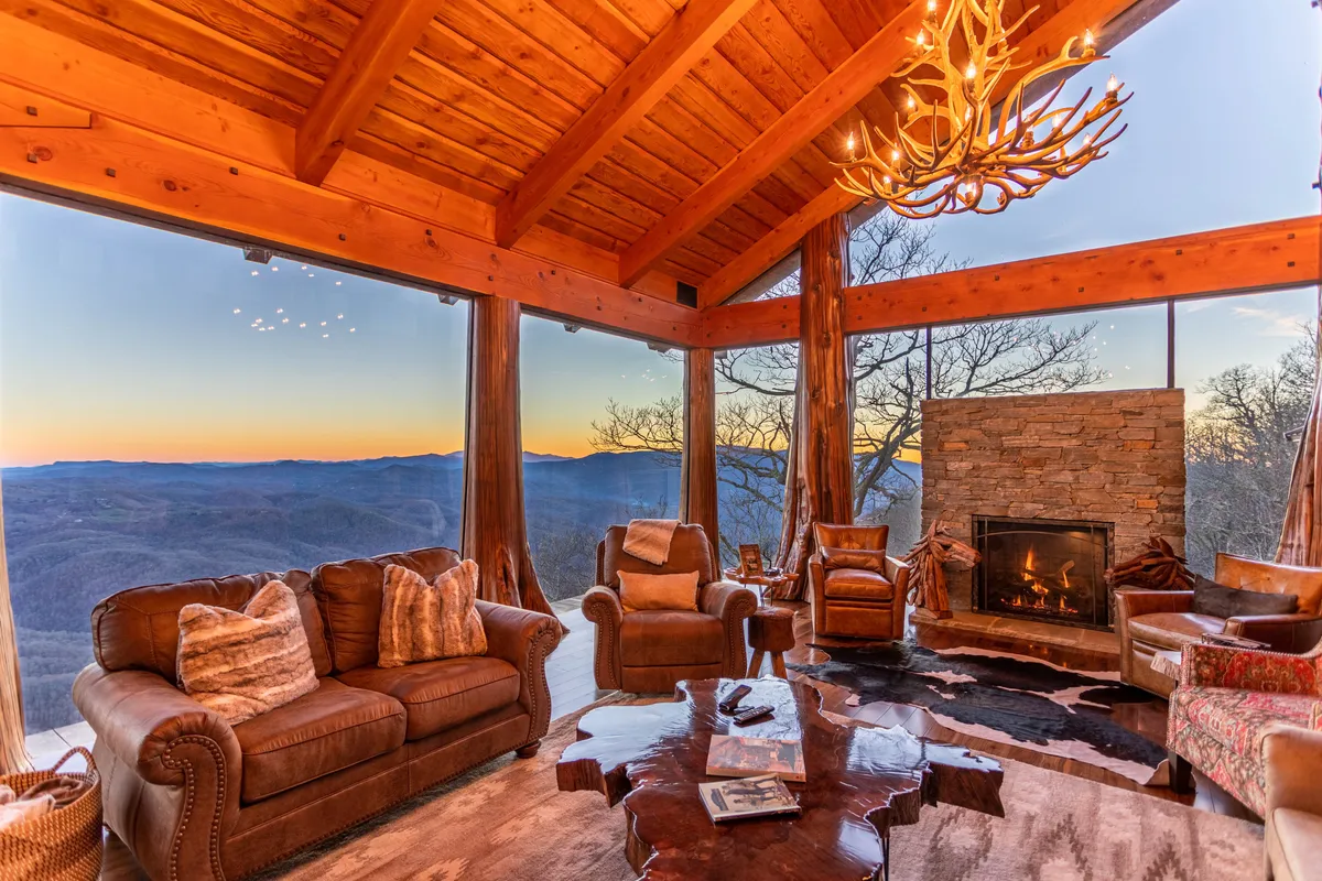 The image captures the luxurious living room of The Flying Eagle, featuring soaring glass walls that frame breathtaking mountain vistas. Inside, a grand stone fireplace and rustic antler chandelier cast a warm glow over plush leather sofas, all set beneath a vaulted wooden ceiling.