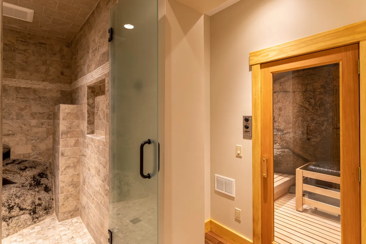 This image spotlights a glass shower enclosure, a wooden sauna door, and textured stone walls, creating a spa-like ambiance. The modern fixtures and overall mood suggest a space for deep relaxation and rejuvenation.