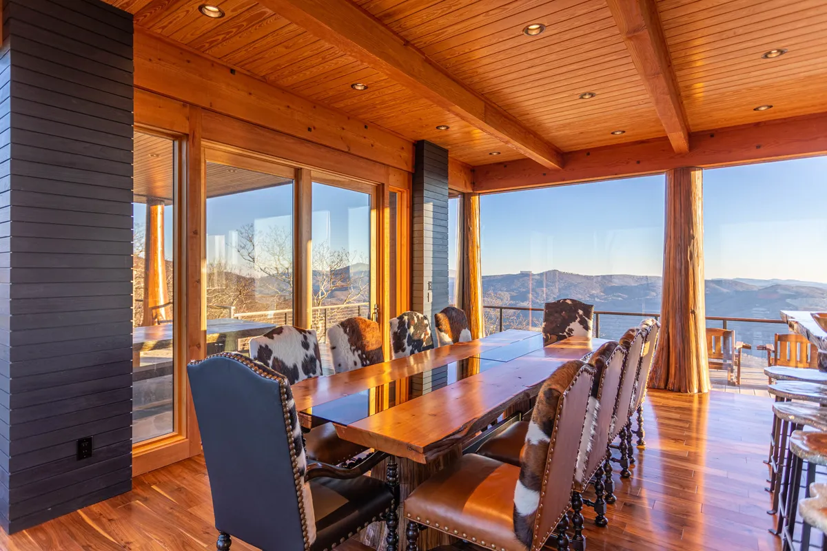 A dramatic dining area is showcased here, featuring a large wooden table surrounded by cowhide chairs and illuminated by floor-to-ceiling windows that reveal sweeping mountain vistas. The setting balances rustic sophistication with a welcoming atmosphere, perfect for group feasts or memorable celebrations.